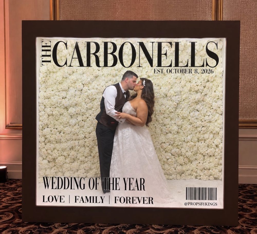 Newlyweds kissing inside a custom magazine booth cover reading The Carbonells, Wedding of the Year, set against a white rose wall